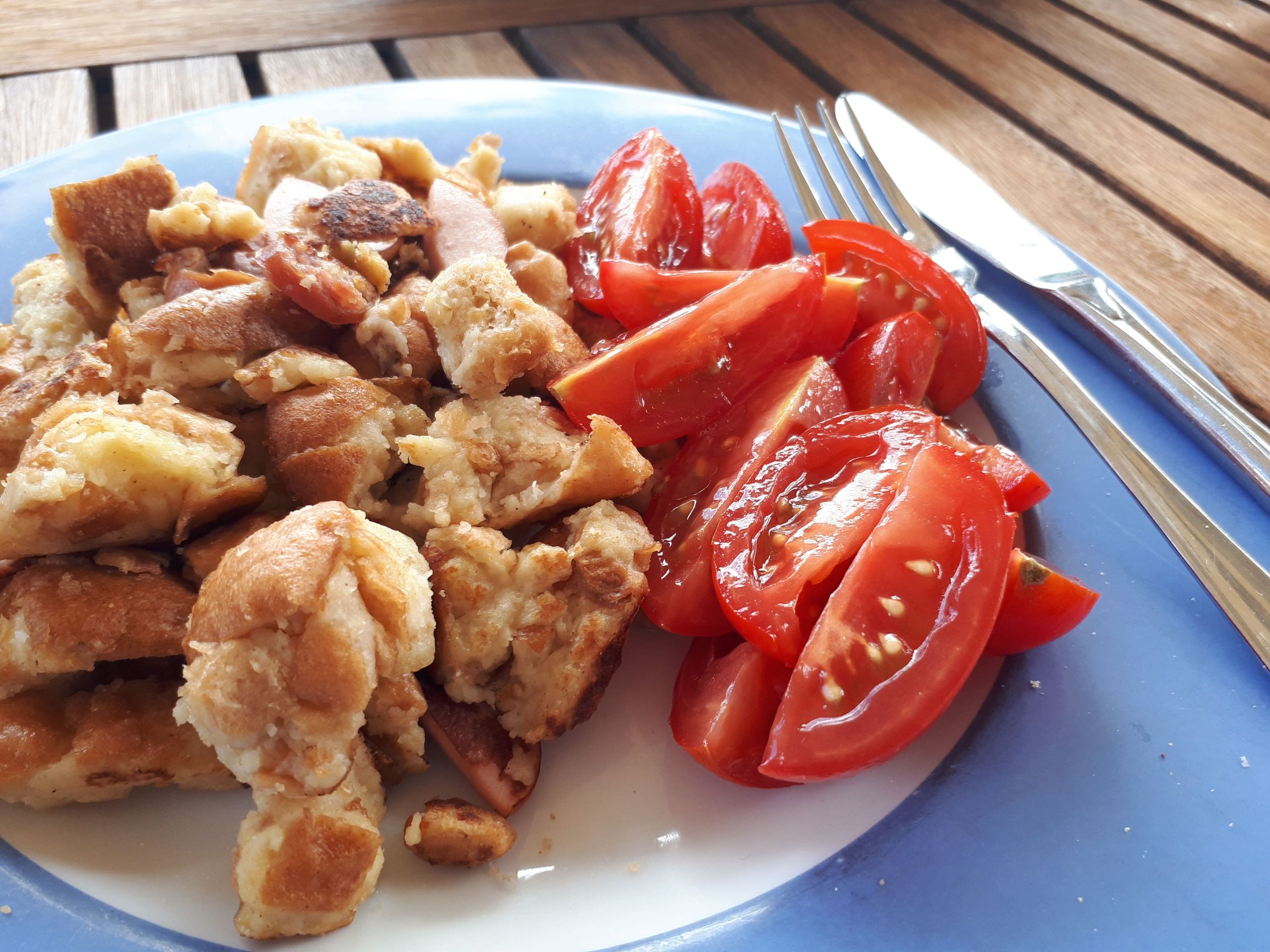 ```   Ein Teller mit zerteiltem Kaiserschmarrn auf der linken Seite und aufgeschnittenen Tomatenstücken auf der rechten Seite. Daneben liegen ein Messer und eine Gabel auf einem Holztisch.   ```